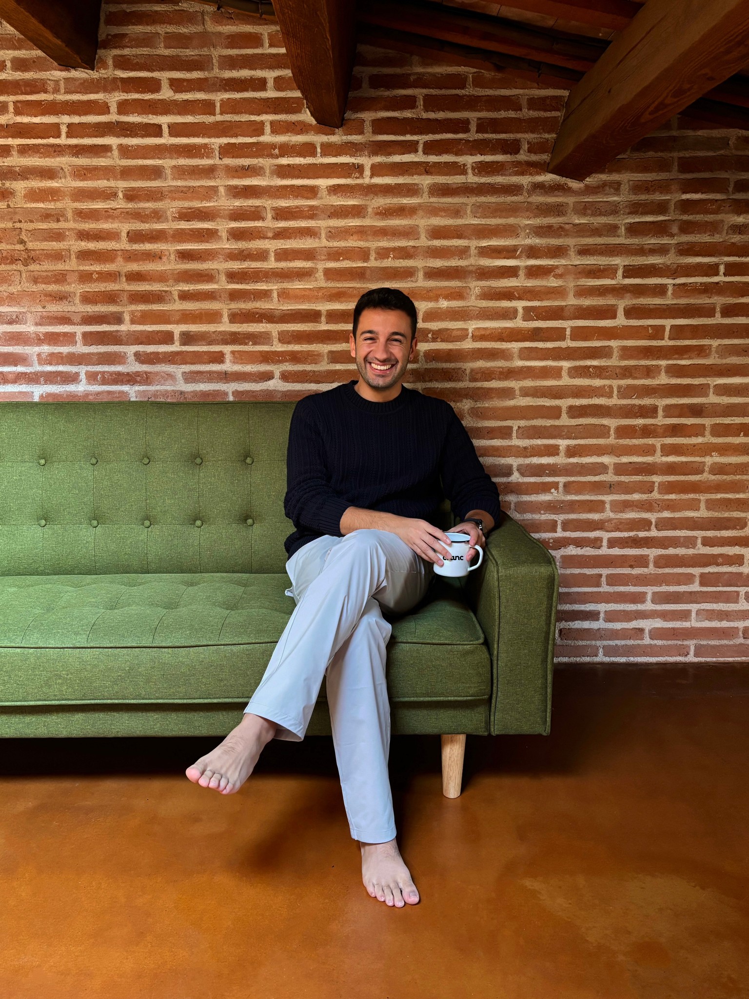 A person sits on a green couch in a cozy room with patterned walls, holding a mug and wearing casual clothing.