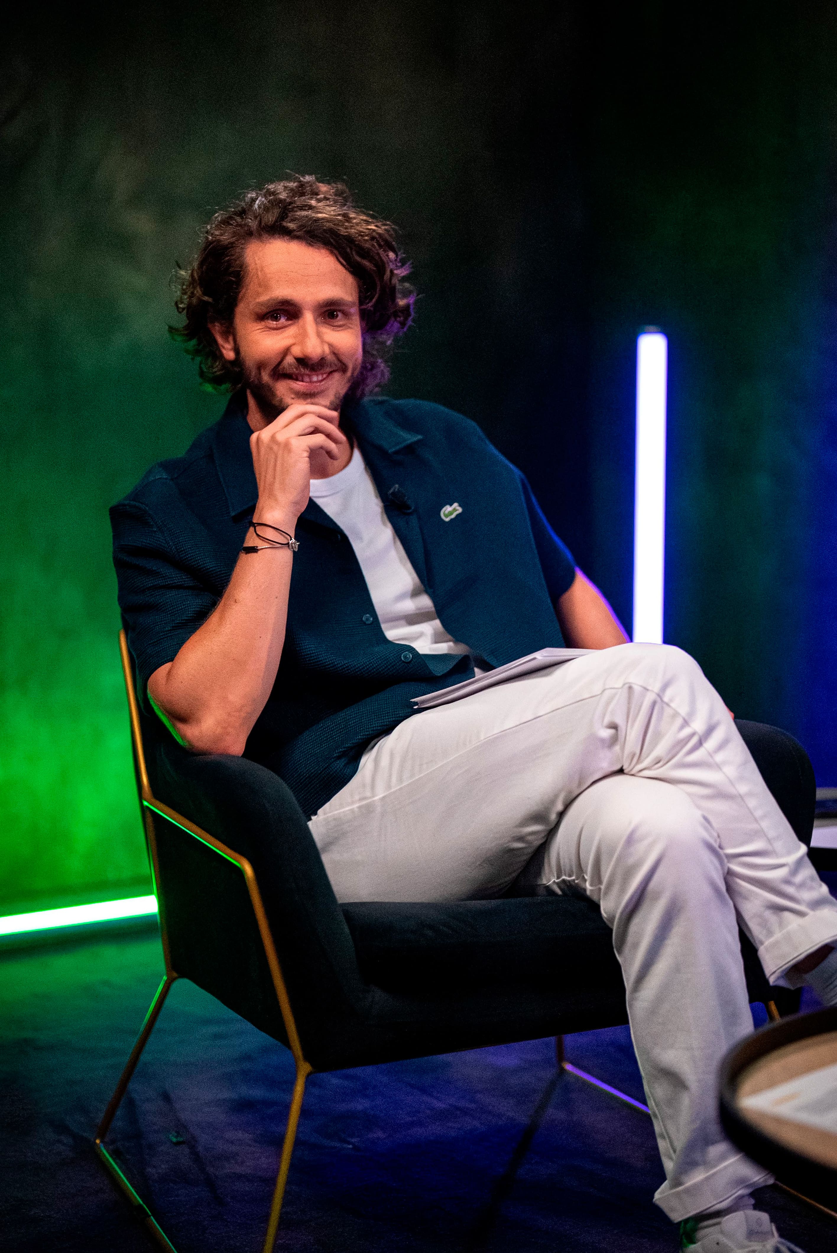 Photographie professionnelle de Guillaume Pley (fondateur Legend) posant sur un fauteuil moderne dans son studio. Il sourit, la main sous le menton, vêtu d'une chemise bleu marine décontractée et d'un pantalon blanc. L'image est caractérisée par un éclairage de studio créatif (lumière néon verte et bleue en arrière-plan), typique des productions de médias numériques. Cette photo de haute qualité, optimisée par retouche et post-production, est un exemple de service Photo & Vidéo fourni par Nux-Studio pour la création de contenu de marque et le branding de célébrités.