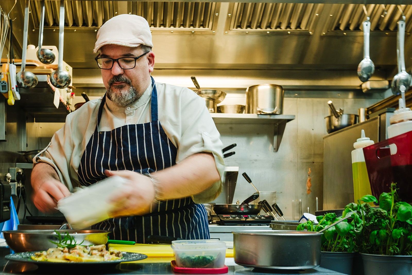 A chef in a striped apron and cap prepares a dish in a professional stainless-steel kitchen, surrounded by fresh herbs, containers of ingredients and cooking equipment, as he works over a counter with focus and precision.