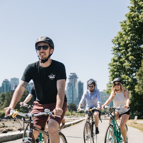 三个人骑着自行车沿着铺设的小路前行,背景是城市天际线和大树。