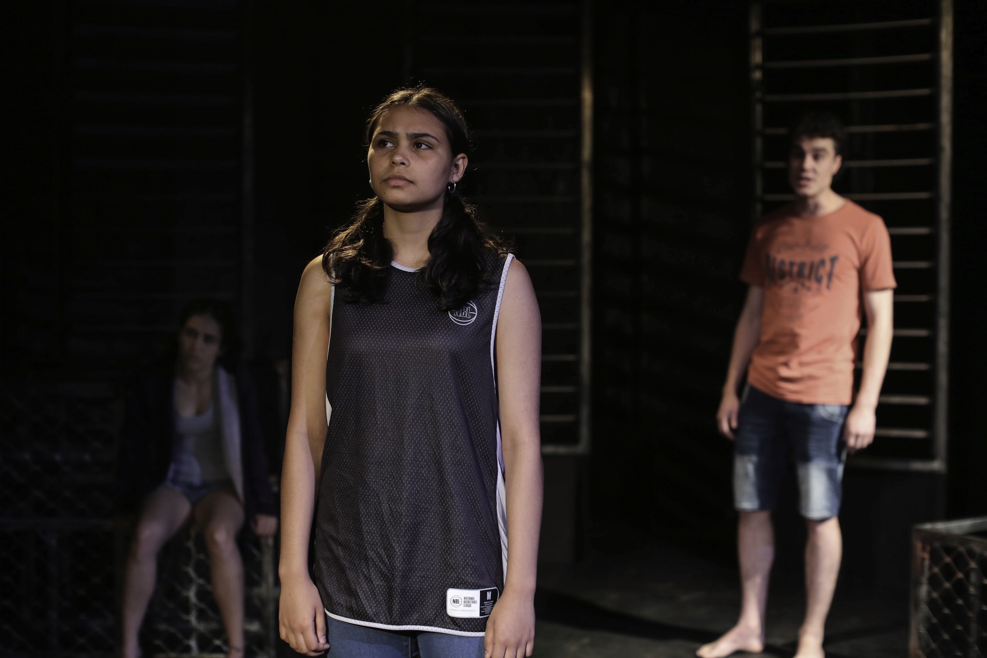 A young woman in a basketball singlet stands centre stage, looking determined. Behind her, a boy in an orange shirt and another girl sit watching in the shadows against a dark, cage-like set.