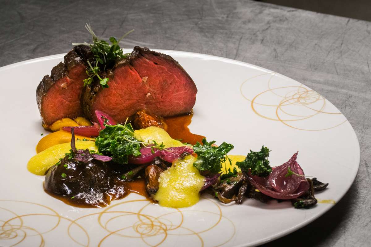 A plated gourmet dish featuring sliced rare roast beef topped with microgreens and a dark reduction sauce on a white plate.