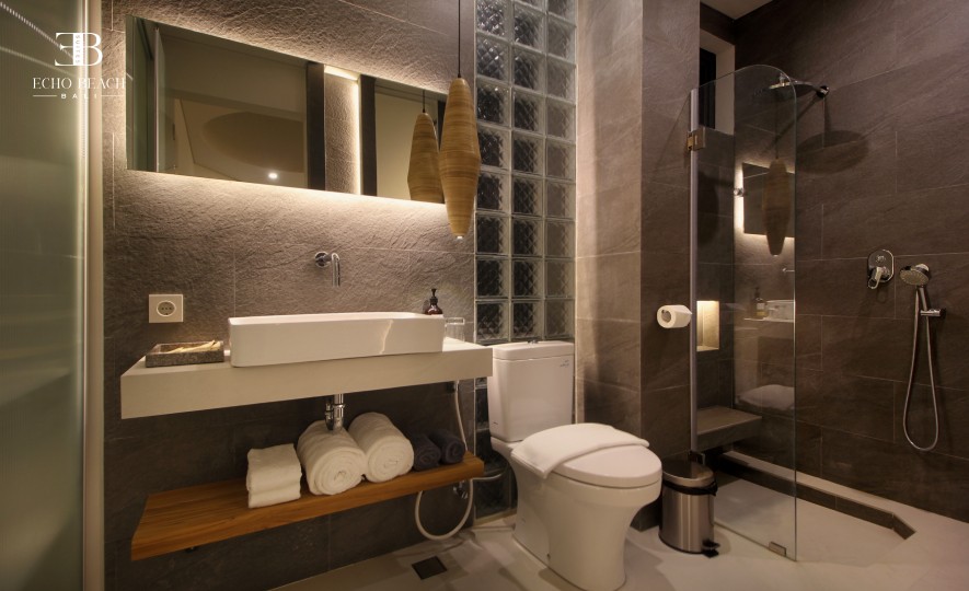 A stylish bathroom at Echo Beach Suite in Canggu, Bali, showcasing modern stone-textured walls, a large backlit mirror, a contemporary vanity, rolled towels, and a glass shower area, offering premium comfort near Echo Beach.
