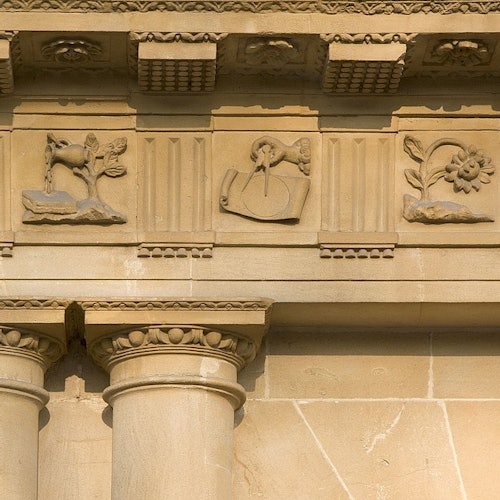 Ornate stone architecture with columns and carvings featuring a goat, a sun dial, and a flower.