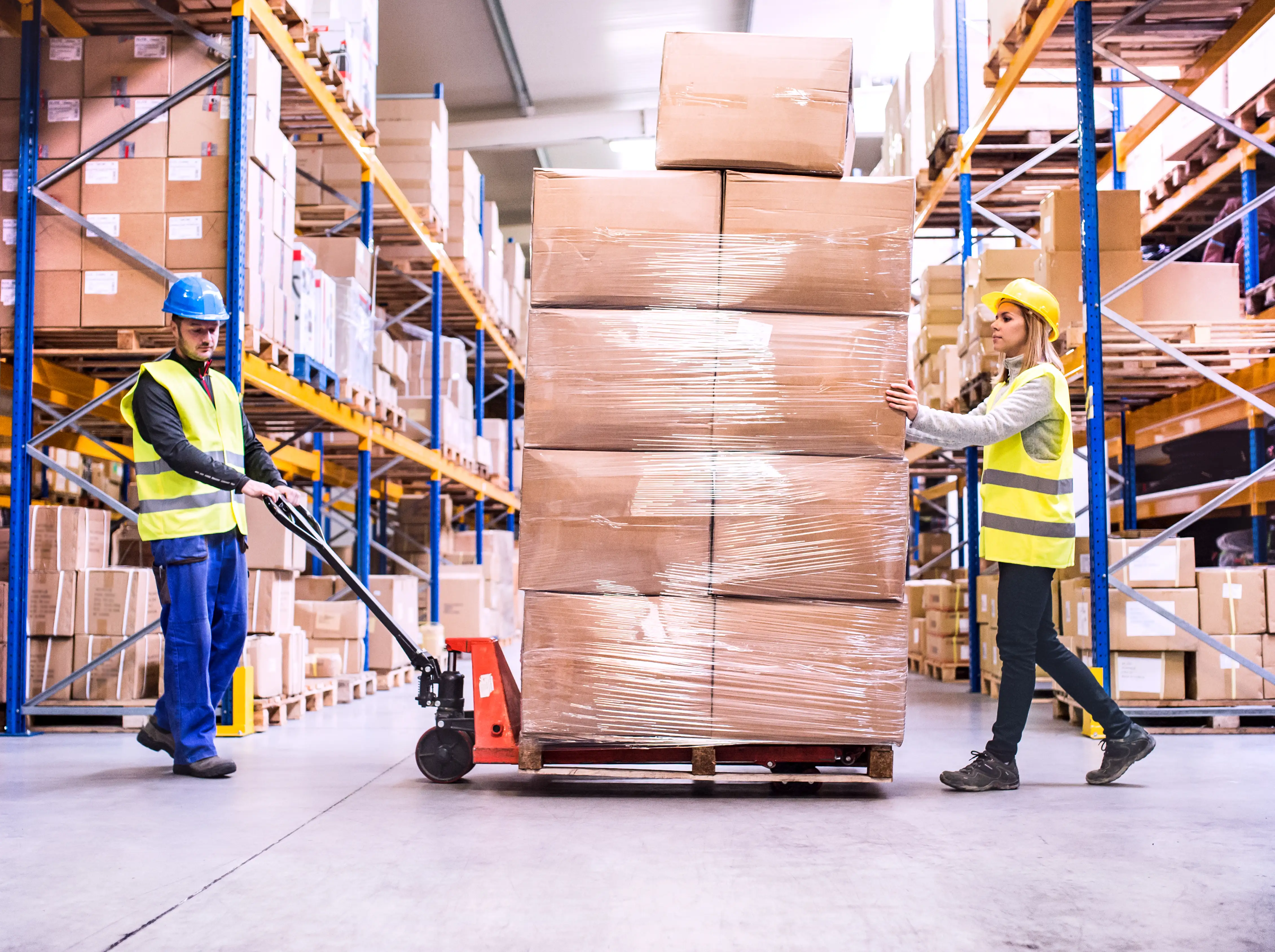 Two warehouse workers moving a large pallet stacked with wrapped cardboard boxes.