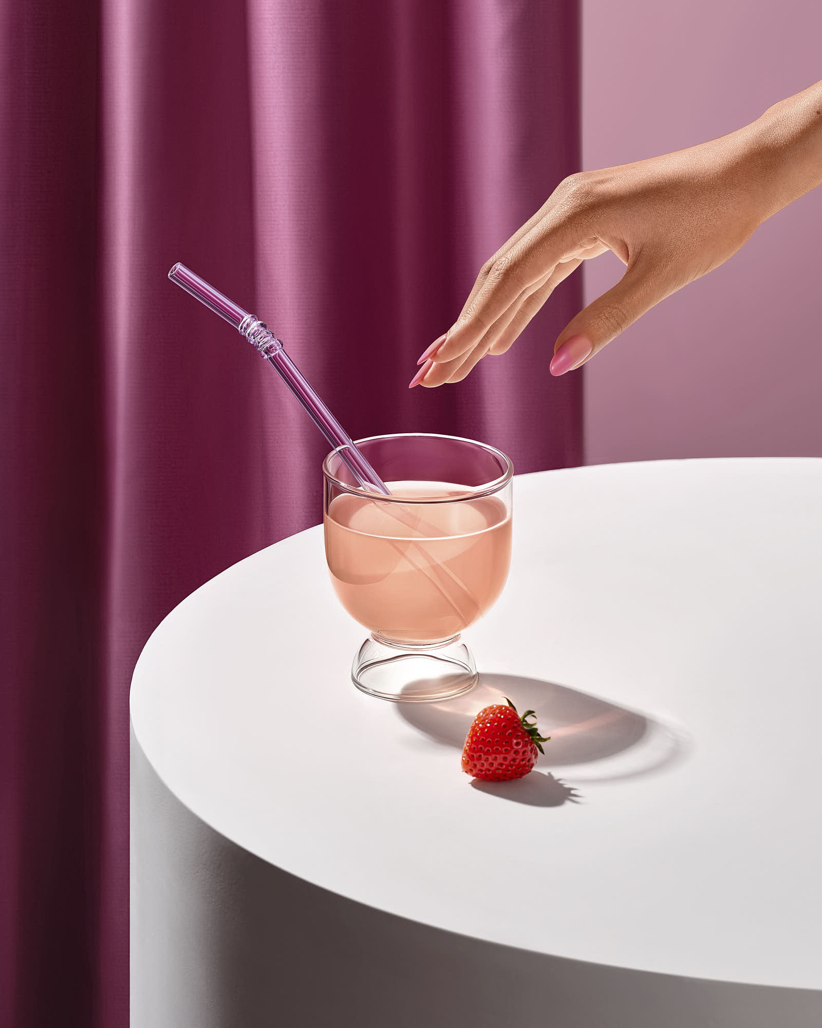 A hand reaches for a pink drink in a glass, with a straw and a fresh raspberry on a white table.