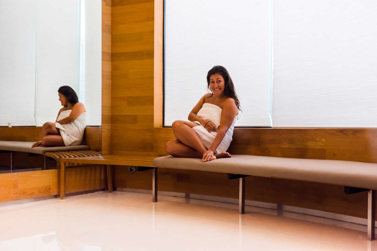 Two women in white swimwear sit on wooden benches in a modern spa with light wood paneling and large windows.