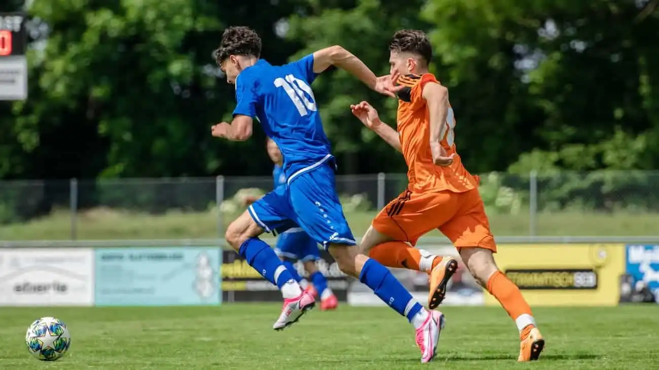 Two football players in a lively friendly match, one in a blue shirt and the other in an orange shirt, fight for the ball on a green pitch, emphasising the importance of selecting suitable opposing teams.