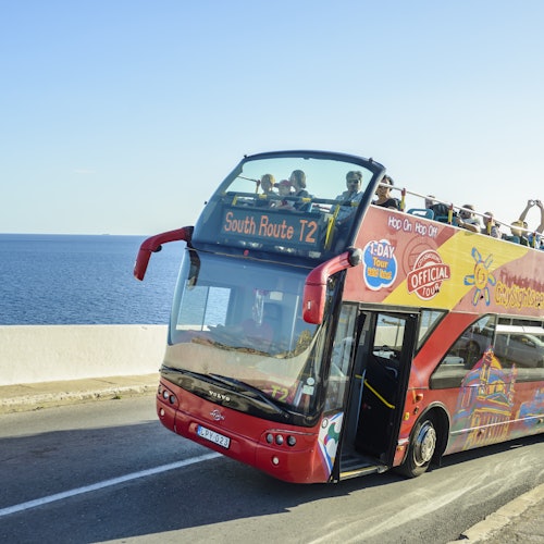 Красный экскурсионный автобус с открытым верхом и двумя этажами, перевозящий пассажиров, едет по прибрежной дороге рядом с спокойным синим морем.