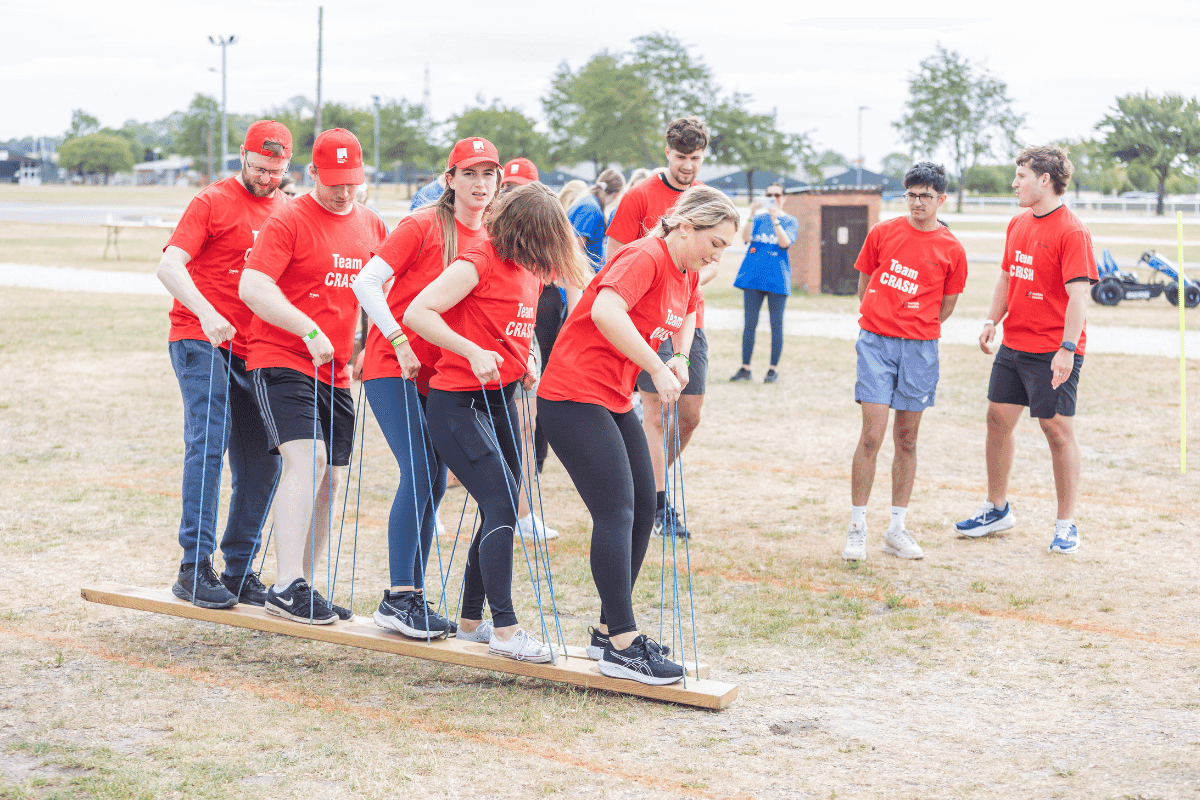 Outdoor team-building event with participants wearing matching blue shirts, taking part in inflatable obstacle courses and games, smiling and cheering, with the words 'ENTER A TEAM' in large bold white and green text across the image.