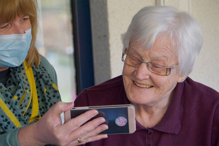 Care worker showing a smartphone to an elderly woman who is smiling.
