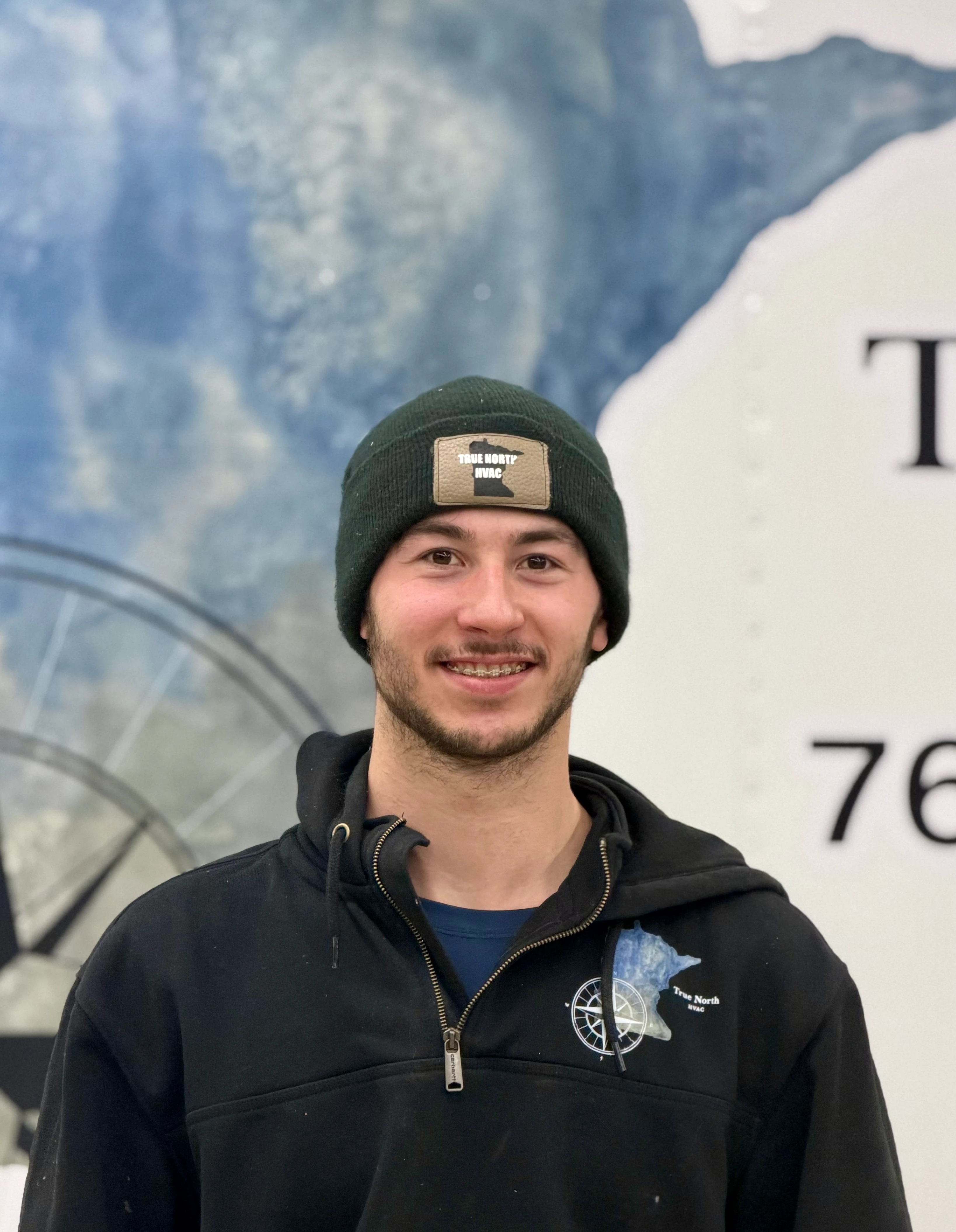Smiling young woman with curly hair sitting at a laptop, in front of a teal wall.