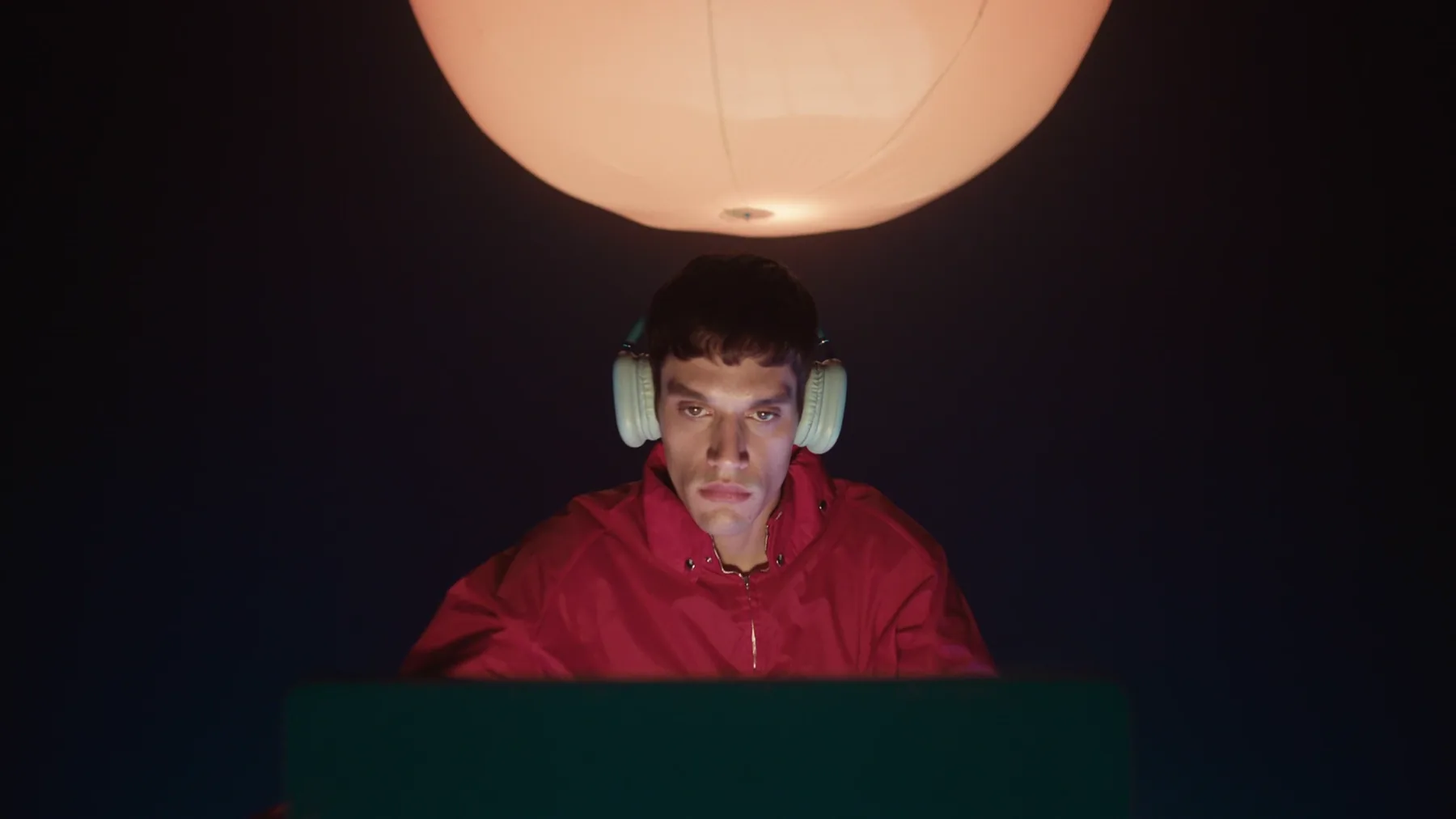Close-up of a young man in a red jacket and pale headphones, face lit from above by a warm orange lamp and from below by the green glow of a laptop.