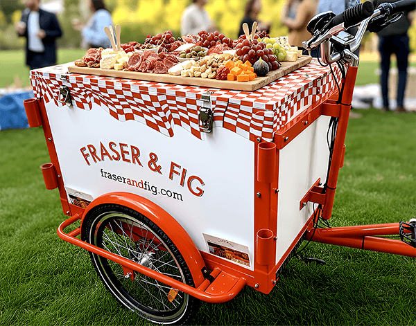 a red-and-white bicycle with fridge box attachment and vinyl wrap on top, reading Fig & Fraser on the sides and front
