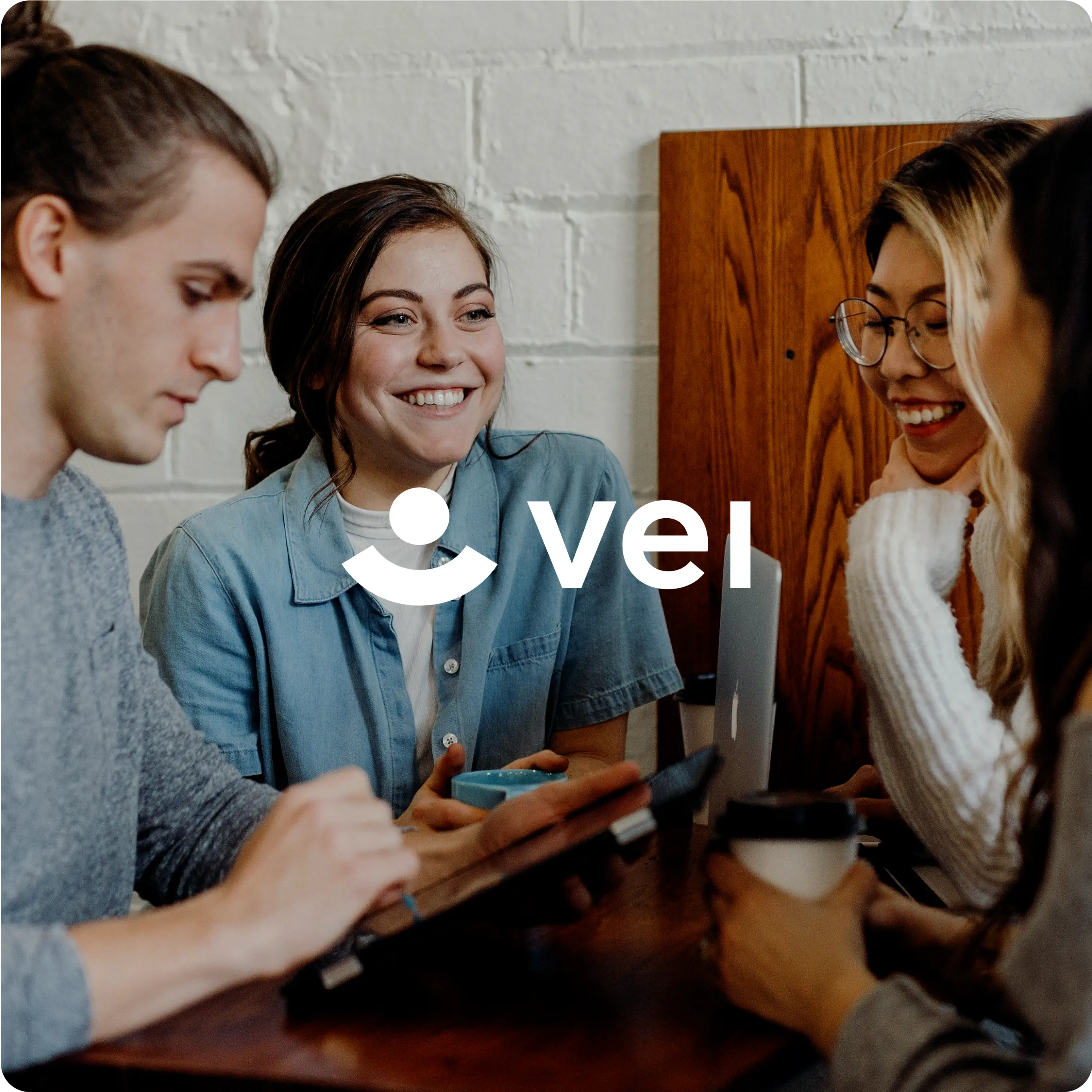 A group of four young adults are gathered around a table, engaging in a lively conversation and smiling. They appear to be in a casual, friendly meeting or study session. One person is using a tablet, another has a laptop, and two are holding coffee cups. The word "vei" is prominently displayed in the center of the image, accompanied by a simple, stylized logo of a person. The background features a white brick wall and wooden furniture, creating a cozy, informal atmosphere.