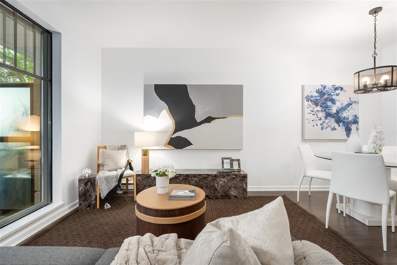 Modern living room with a large window, light-colored furniture, a round coffee table, and decorative artwork.