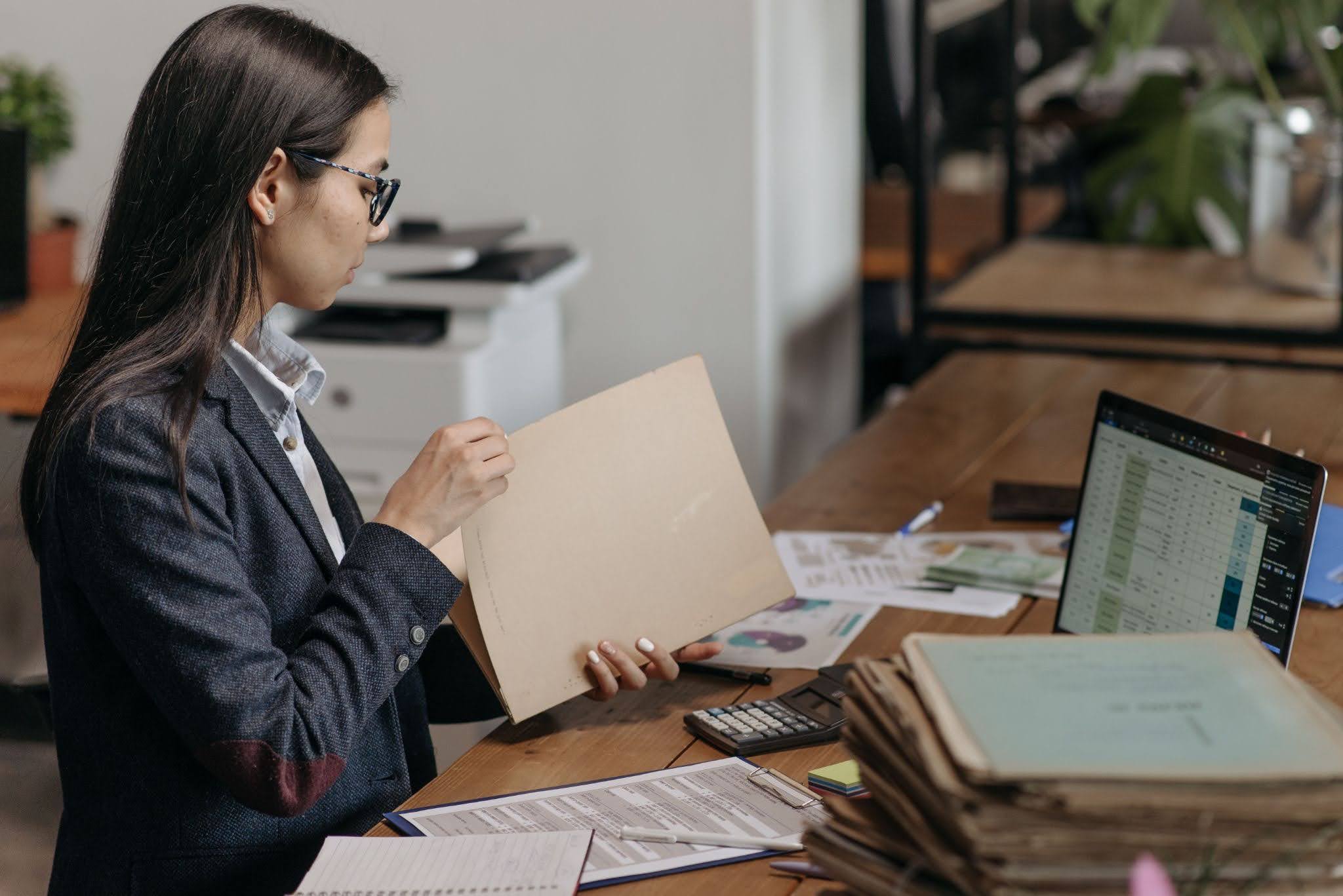 Professional woman organizing files at desk with laptop and documents piled up. 