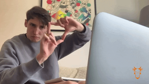 A man in a gray sweatshirt sits at a laptop and forms a heart shape with his hands toward the screen.