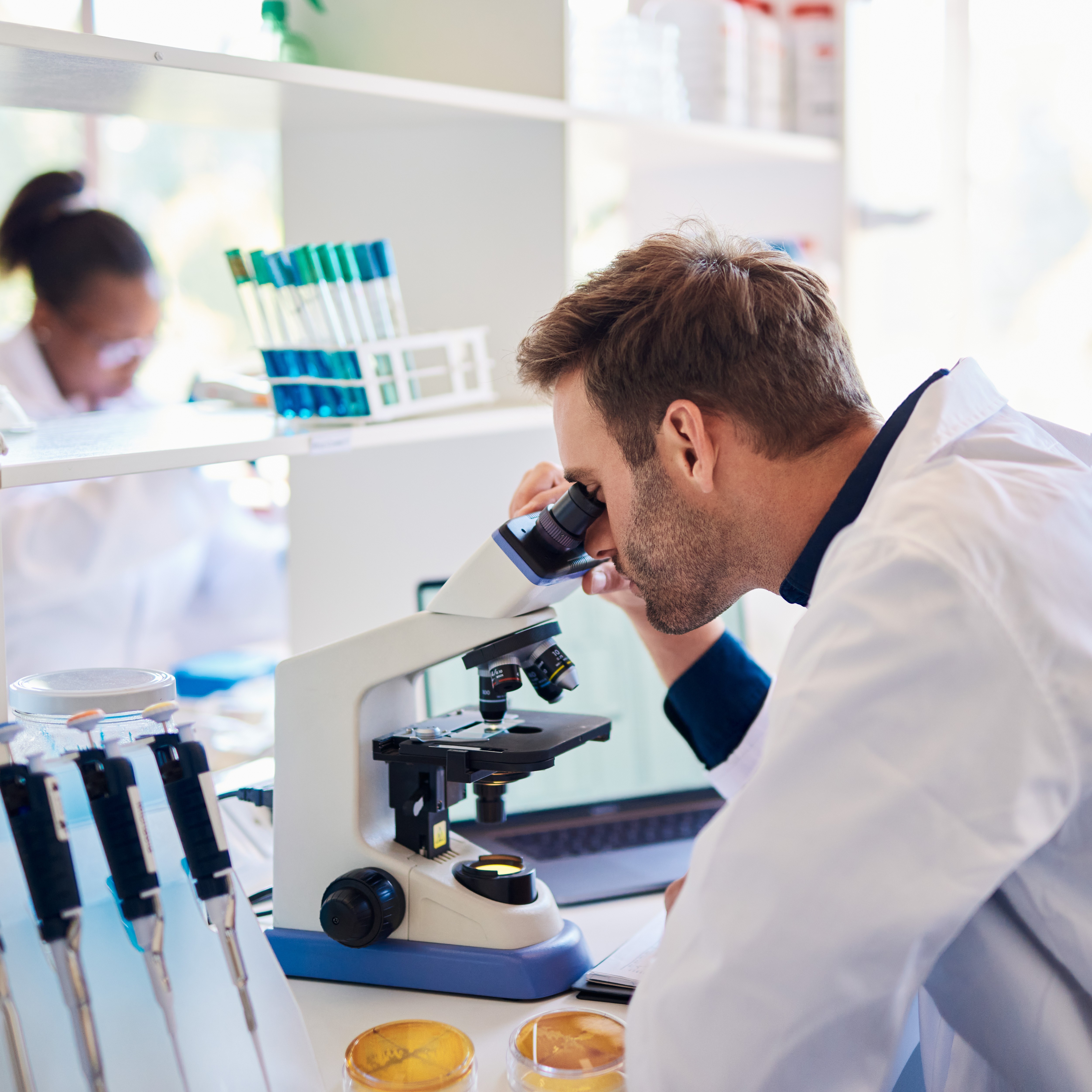 Scientist in a lab coat examining samples under a microscope in a pharmaceutical research laboratory, demonstrating the specialized talent necessary for medical innovation and drug development.