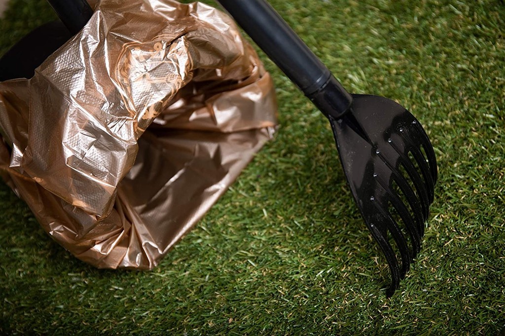 A garden spade leans against a brown bag on green grass, ready for scooping poop.