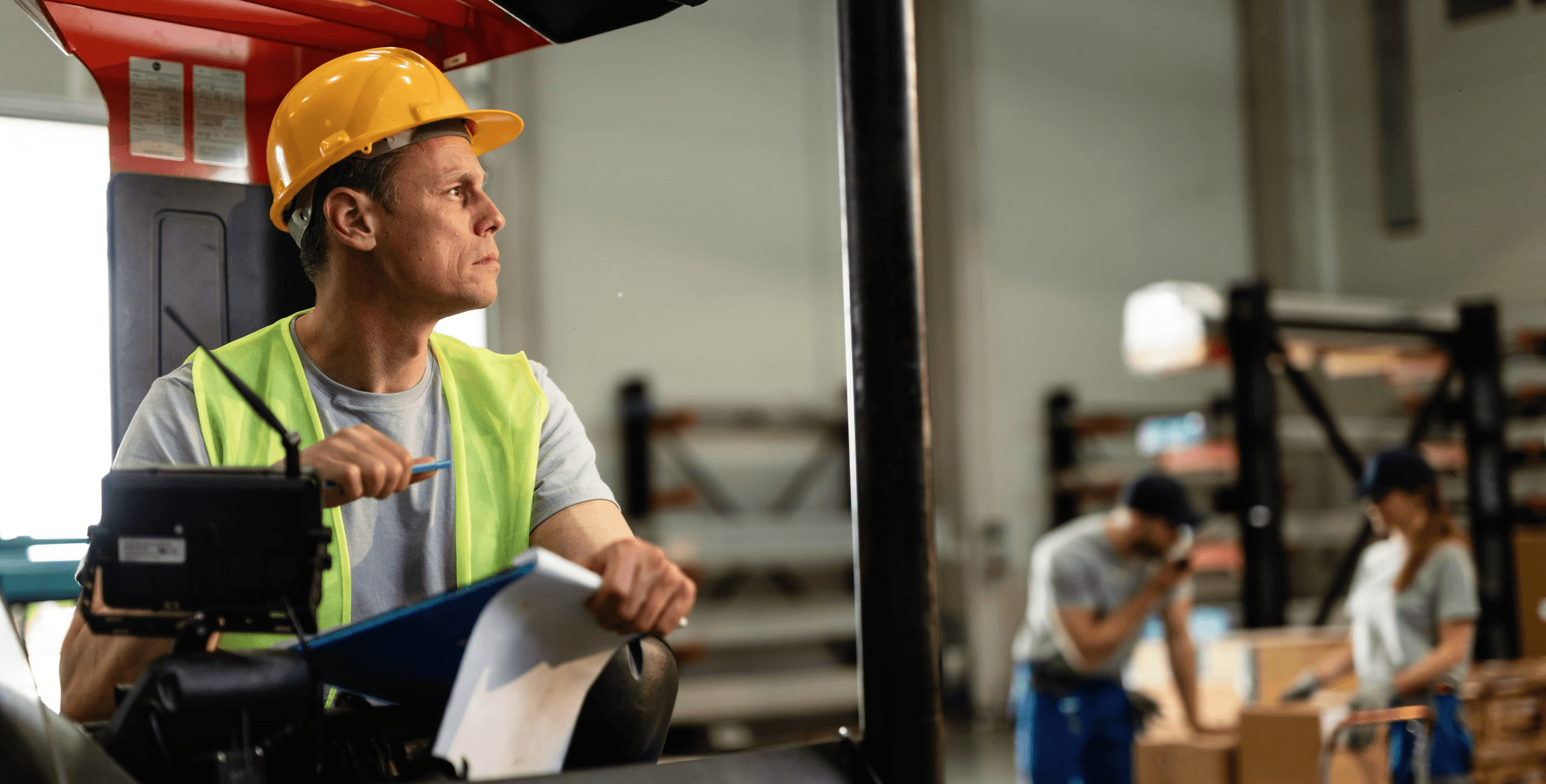A warehouse worker in a safety vest and hard hat operates a forklift while holding a clipboard, with colleagues organizing boxes in the background.