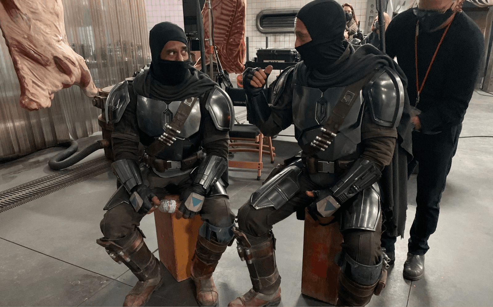 Two individuals in tactical gear sit on wooden boxes, engaged in conversation while a third person stands nearby.