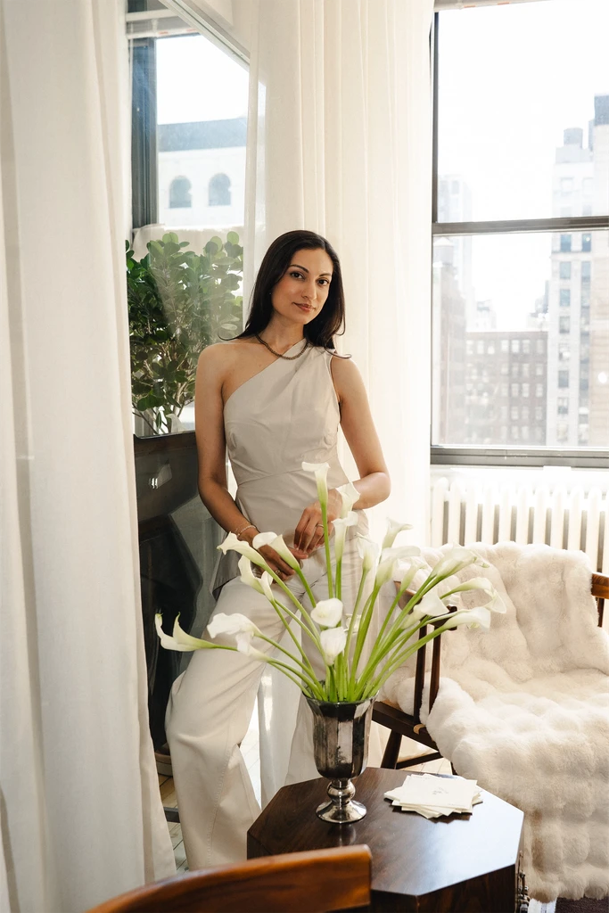Saher Hussaini, founder of SYI Acupuncture, standing by a sunlit window beside white calla lilies