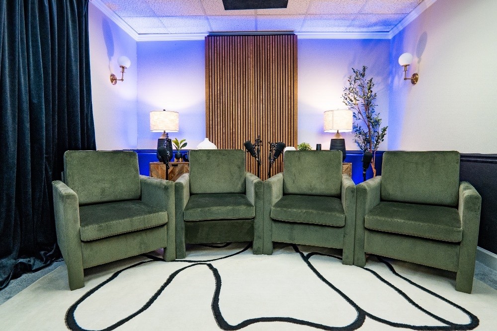 Book Studio A “Studio A setup with four green armchairs arranged in a semi-circle on a white rug with black abstract lines, framed by dark curtains and warm wood paneling under purple ambient lighting.”