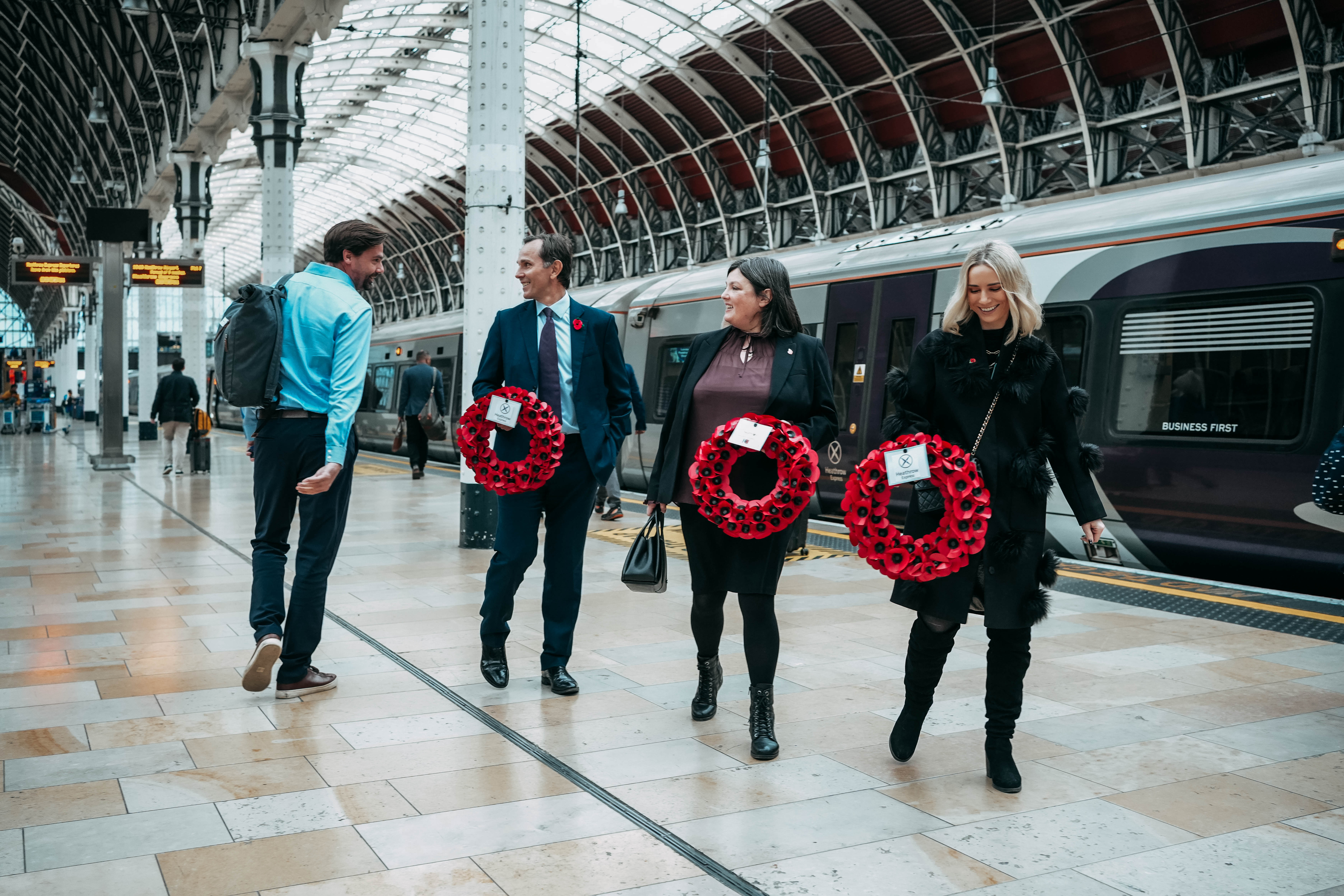 Four people, three of whom are carrying large red poppy wreaths, walk and chat along a platform in a spacious, modern train station next to a stopped train.