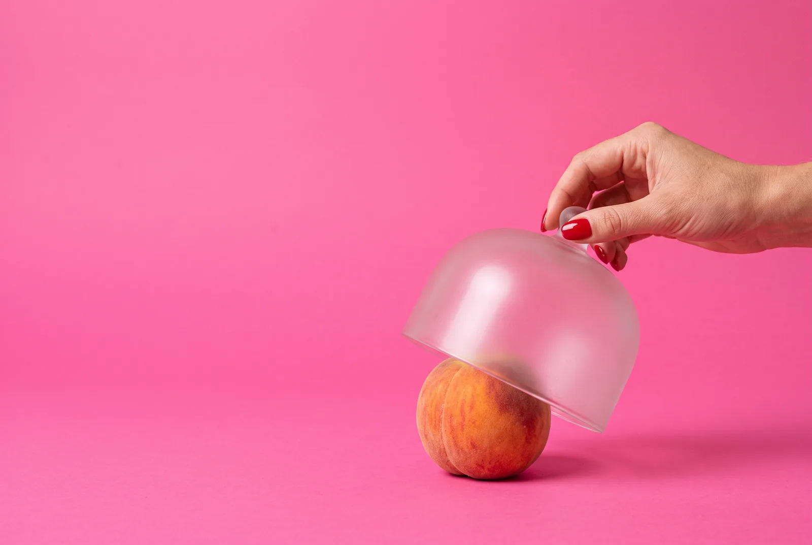 A hand with red nails lifting a frosted glass cloche to peek at a hidden peach, showing why a secret AI girlfriend can feel like cheating