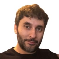 A person with short, wavy dark hair, a beard, and mustache is wearing a black shirt against a plain white background.