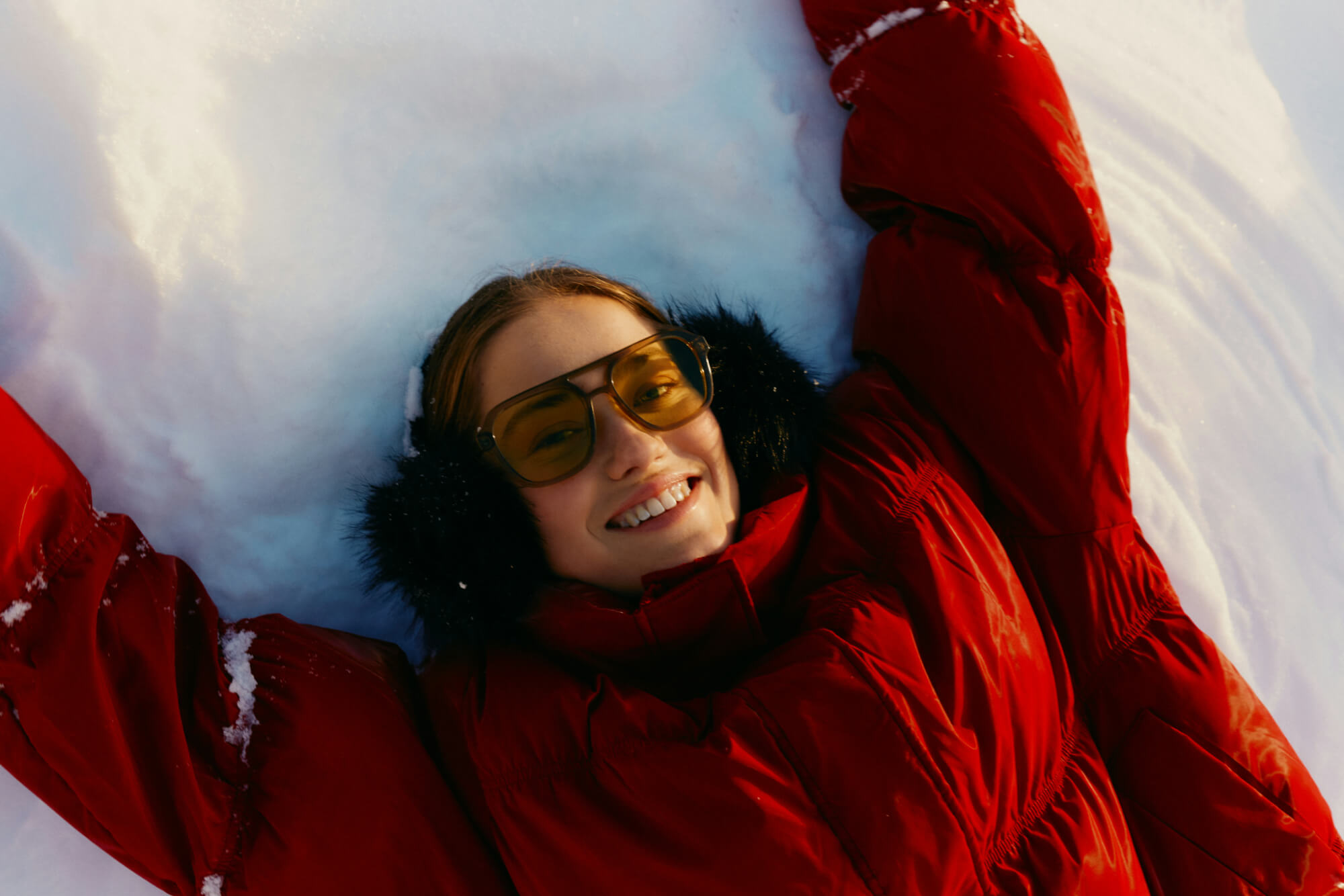 A person in a red coat with earmuffs and large sunglasses smiles while lying on snow, arms outstretched, conveying joy and warmth in a winter setting.