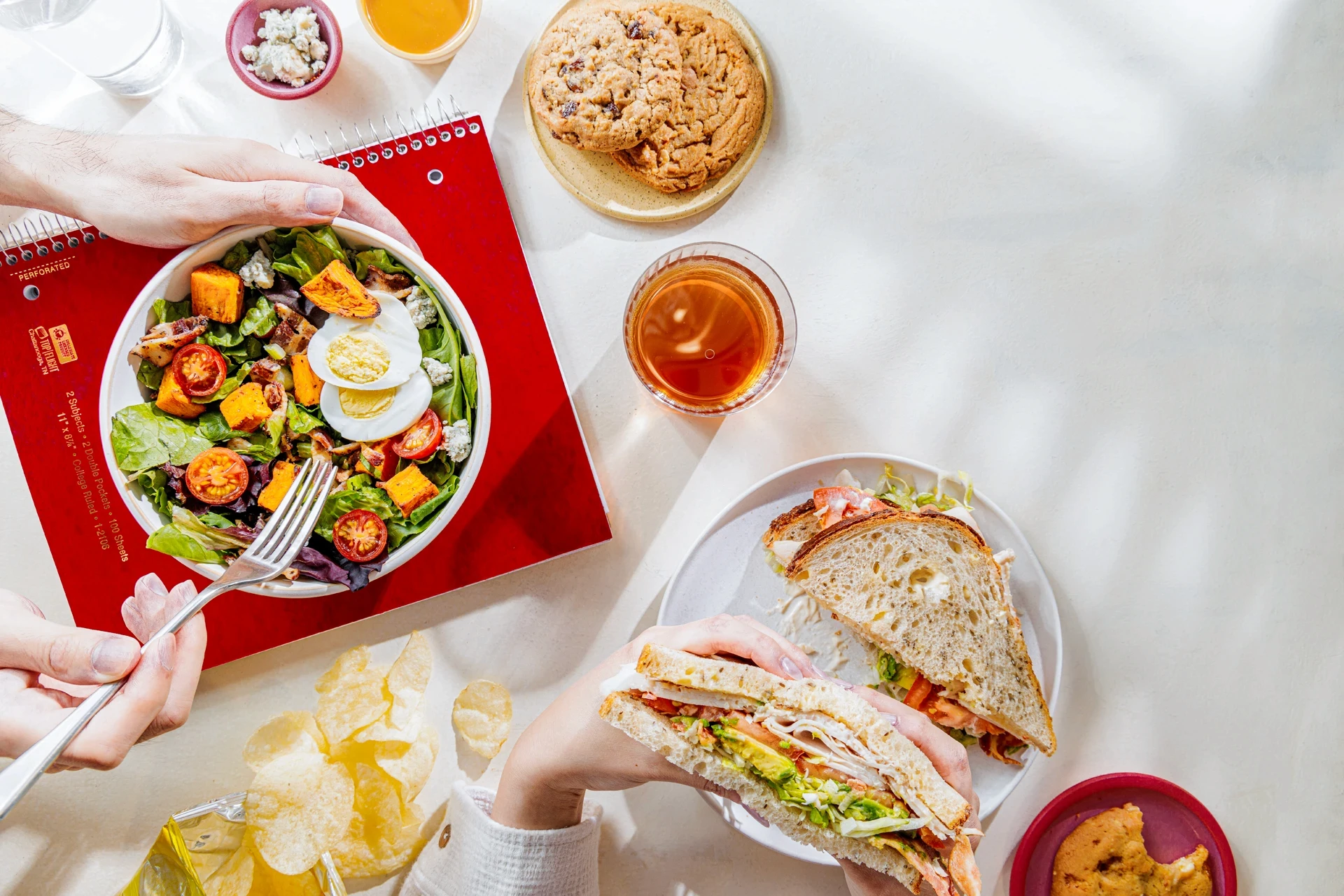 Top view of people's hands eating a salad bowl and sandwich.