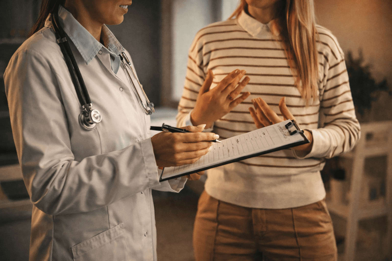 Person in white coat with stethoscope writes on clipboard for another person.