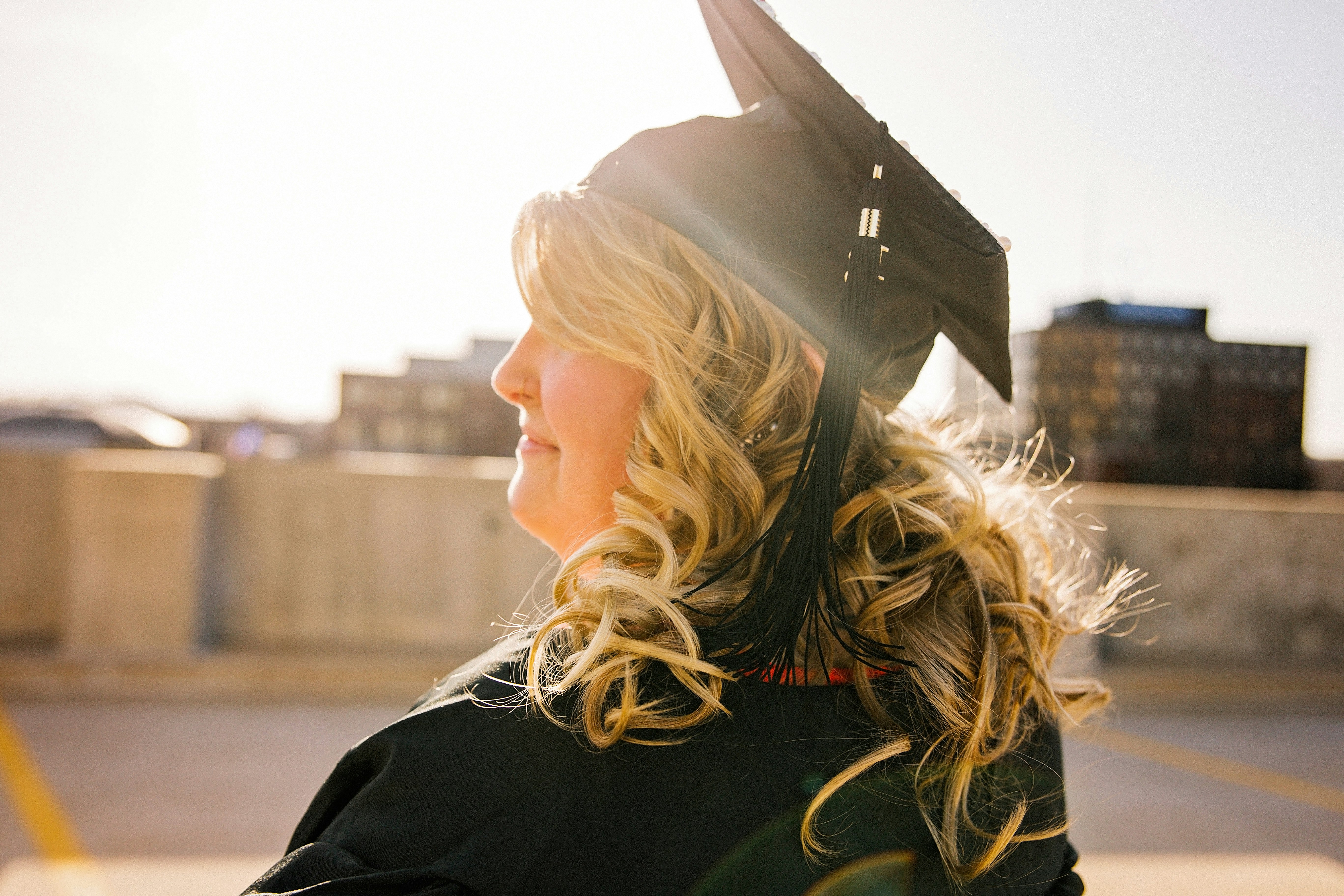 A woman wearing a graduaion cap and gown stands facing the left, in direct sunlight. She has long, blonde, curly hair. Behind her buildings can be seen in the distance across from a parking lot.