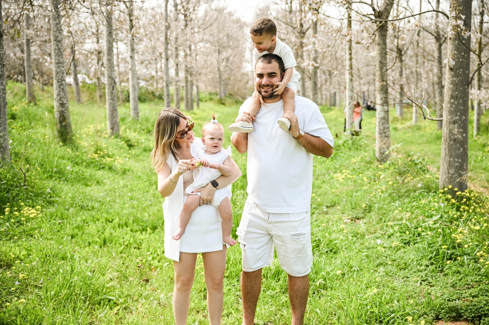 פורטרט משפחתי מלא חום בלב חורשה פורחת. זוג הורים מחייכים ומביטים בילדיהם הקטנים באווירה אביבית ונינוחה תחת אור טבעי, מאת Shiriz Photography הצלמת שירי עציץ.