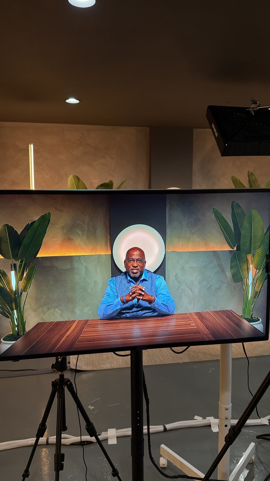 A presenter at a wooden desk, framed by warm lighting and greenery on the production monitor.