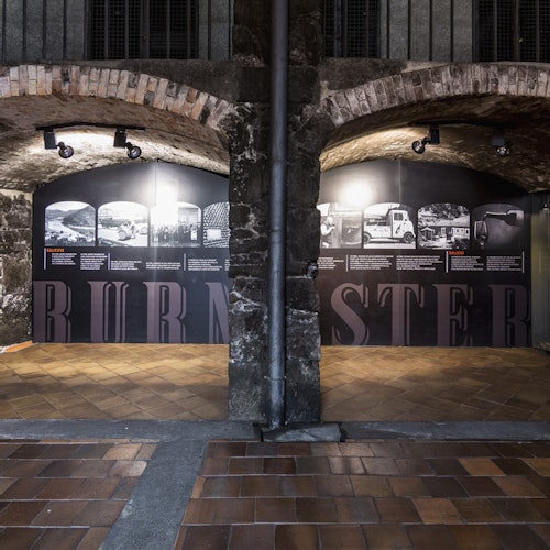 A stone archway with tiled flooring leads to a museum display featuring historical photographs and text on black walls.