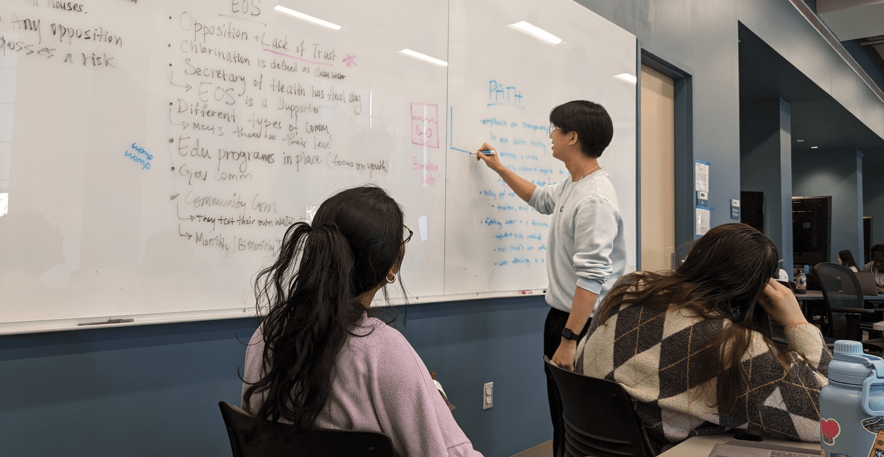 Huy stands at a whiteboard, writing notes. Anagha and Stacie look on.