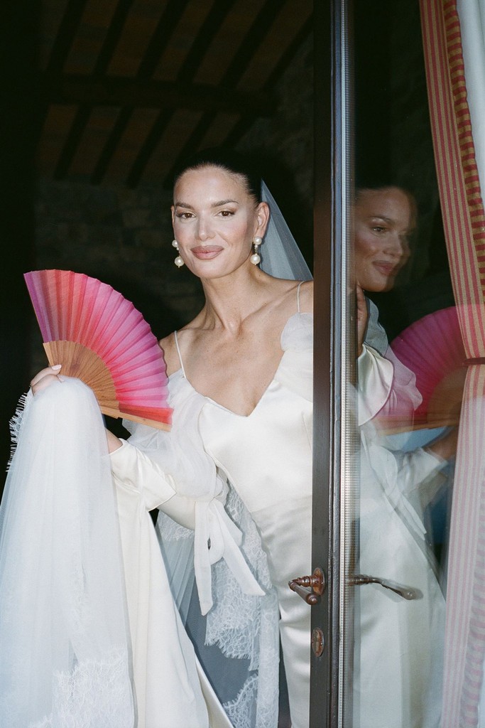 Bride in a silk wedding dress holding a pink fan while standing by a door