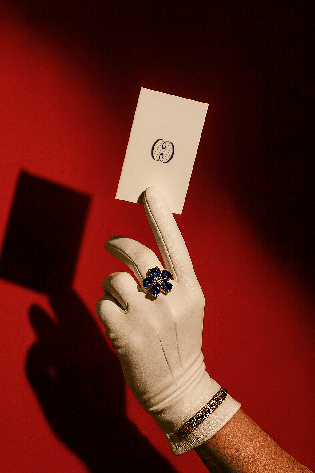 Gloved hand holding a cream card with a logo, wearing a gold bracelet and a blue gemstone flower ring against a red background.
