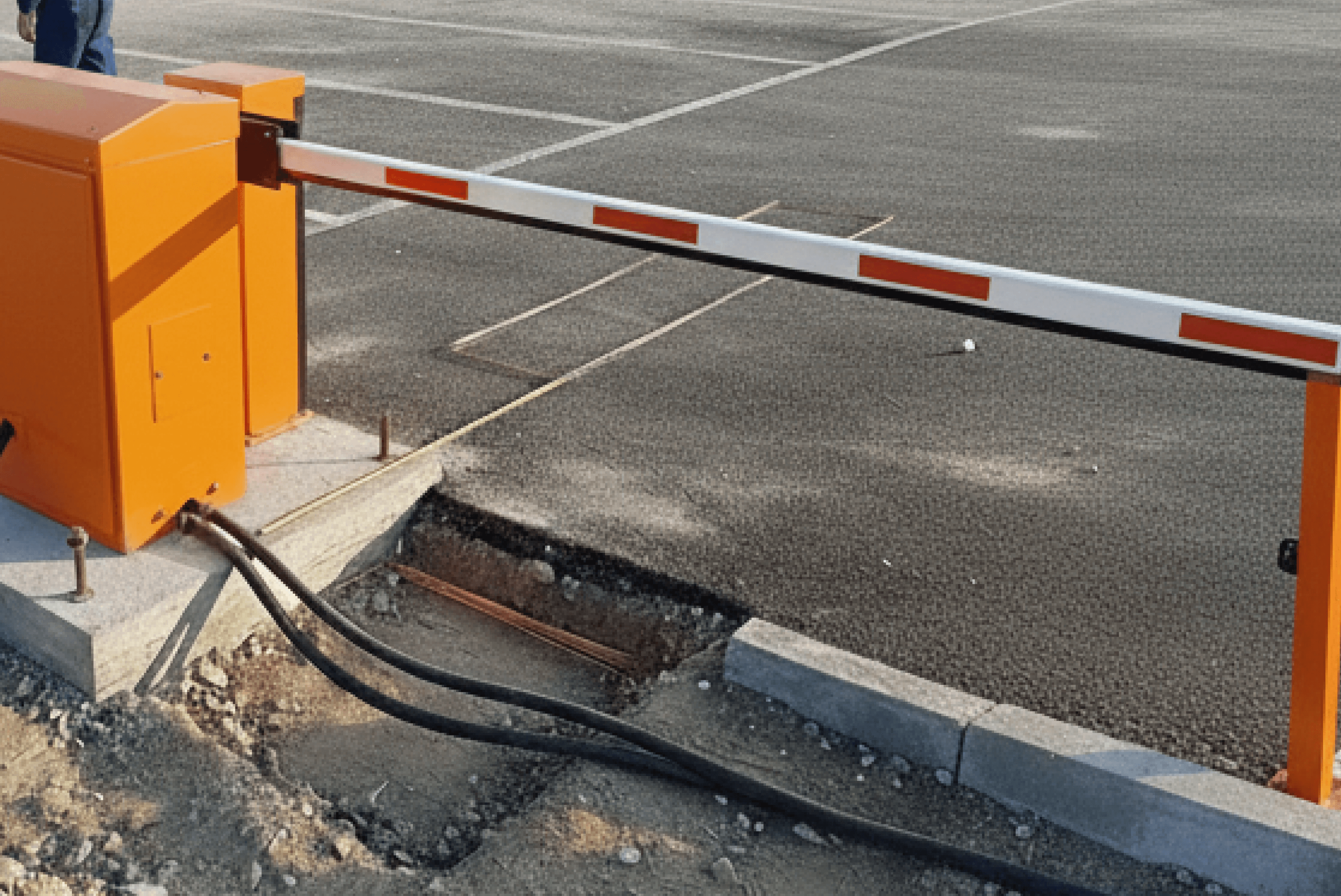 야외 주차장 입구에 오렌지색 차단기 본체와 바(bar)가 설치되어 있으며, 하부에 배선 작업을 위한 케이블과 굴착 흔적이 보임. 시공 진행 중인 현장 모습입니다.