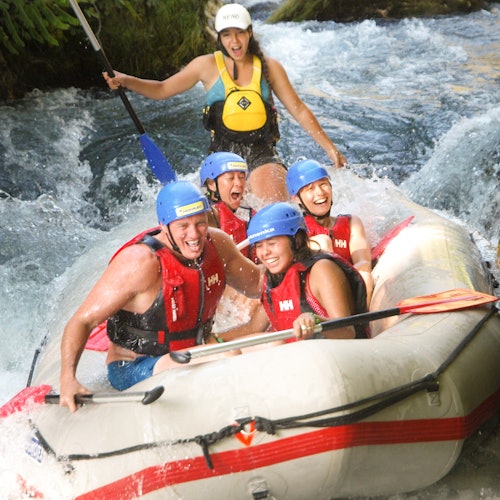 Fyra personer i flytvästar och hjälmar åker glatt nedför en flod, med en instruktör bakom dem som navigerar det turbulenta vattnet.