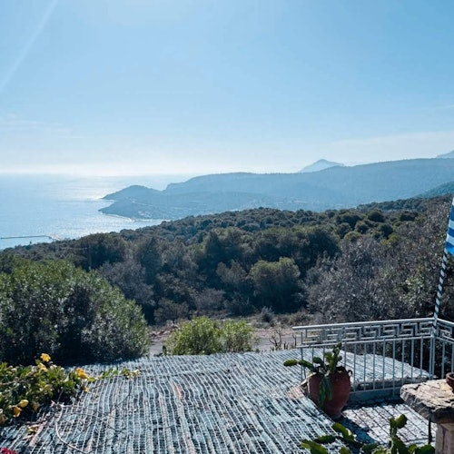 一幅风景优美的海岸景观，远处是连绵的山丘、平静的海面，以及在晴朗蓝天下摆放着盆栽植物的露台。