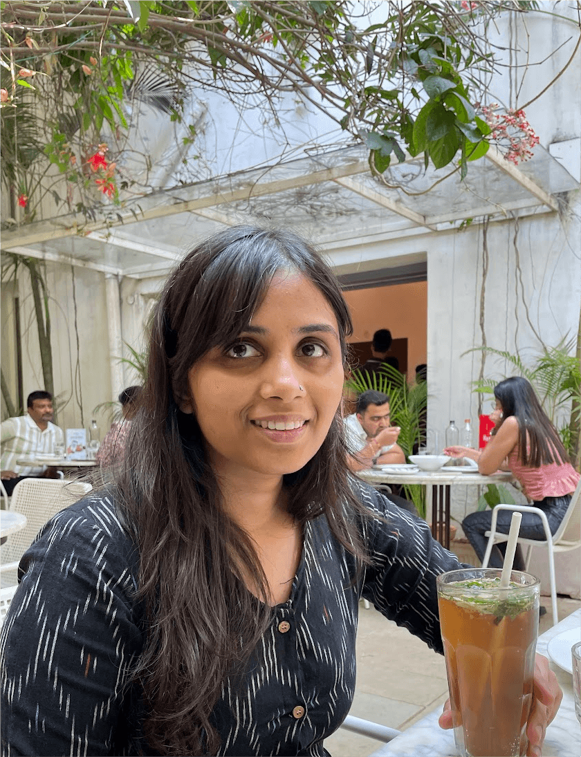 A photo of Prajna Nayak smiling, with a drink in hand. There's a cafe in the backdrop, and people sitting at tables.