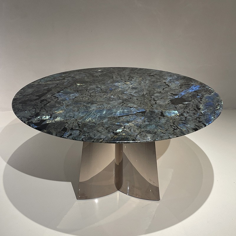 A round table with a polished labradorite stone surface, featuring a dark bluish-gray color with reflective, iridescent patches. The table stands on a sleek, metallic base.