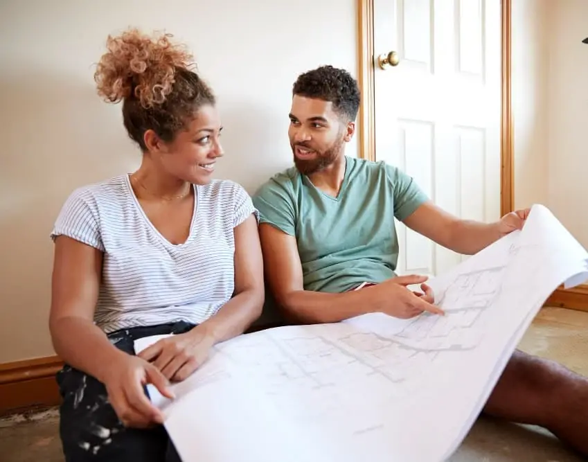 A couple sits on the floor of their new home, smiling and reviewing blueprints together. The image represents the excitement and planning that come with building a dream home through Chris Lewis Home Loans.