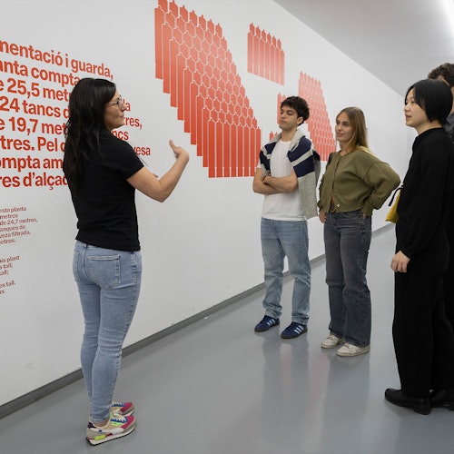 一位女士在对三位女性和一位男性讲话时做出手势，墙后的白色墙壁上有红色文本和图形。