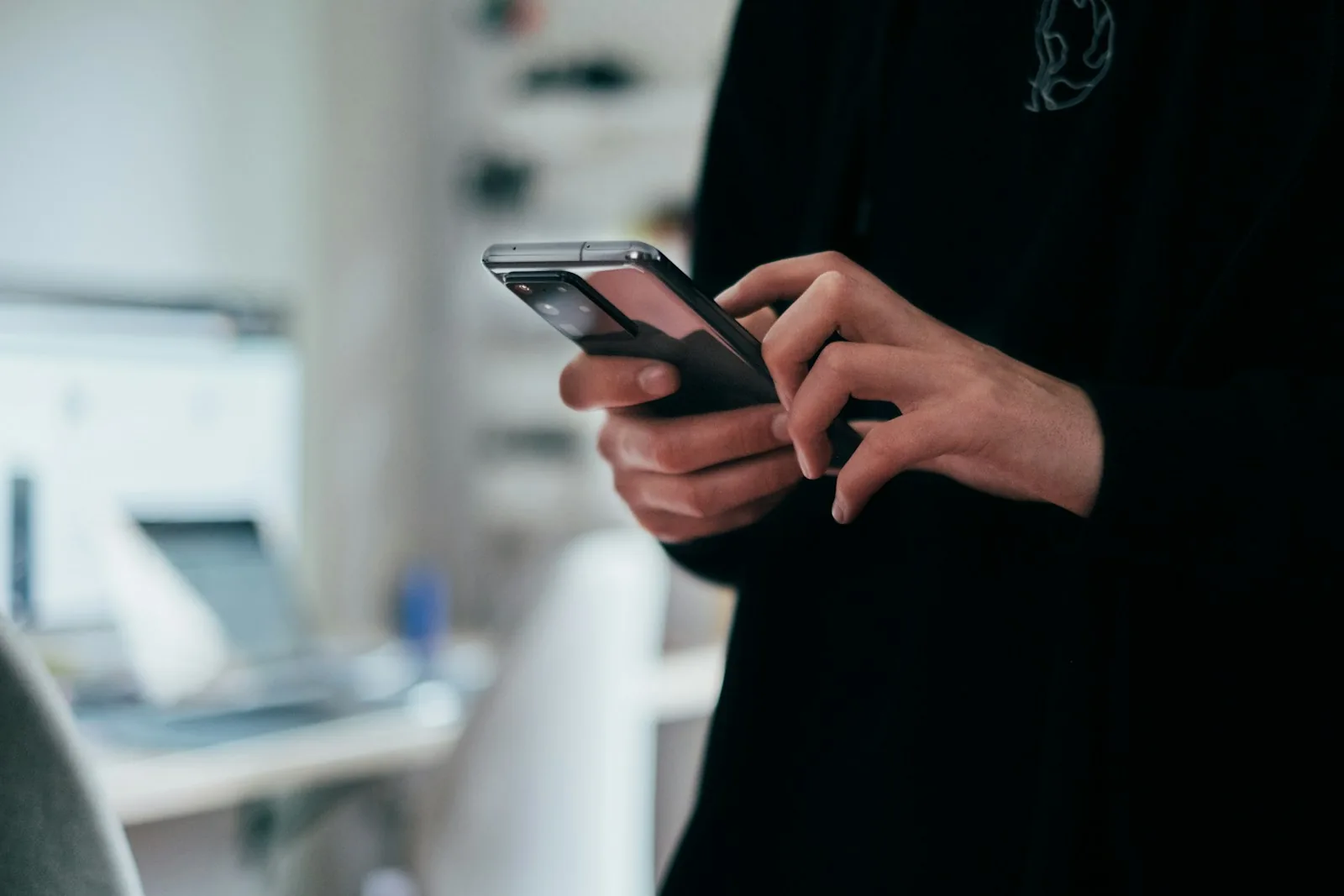 Close-up of two hands holding and tapping a smartphone, with a blurred desk, laptop and monitor in the background