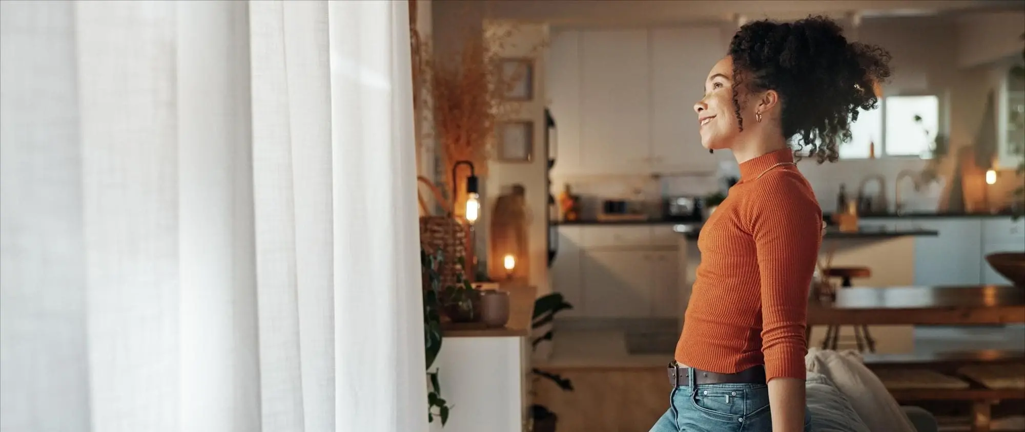 A woman stands by a bright window, smiling as sunlight fills her cozy, modern kitchen and dining area. The image conveys optimism, warmth, and the confidence that comes from successful real estate investing with Chris Lewis Home Loans.